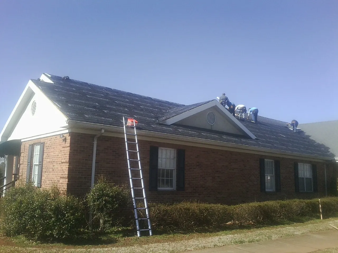 Crew tearing shingles off a brick ranch roof with a ladder set — Metal Roof Repair work by our crew in Inwood